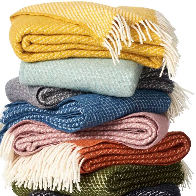 Mehrere gefaltete Decken der Serie Velvet, übereinandergestapelt, in verschiedenen Farben wie Gelb, Blau, Grau, Rosa, Orange und Grün, mit weißen Fransen an den Rändern. Die Szene vermittelt ein Gefühl von Gemütlichkeit und Wärme sowie Auswahl an modernen Wohntextilien.