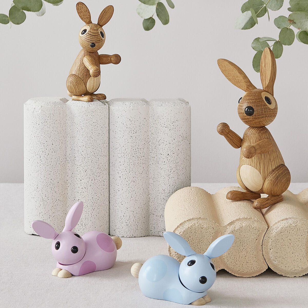 Zwei sitzende Holzhasen und zwei liegende Hasenfiguren in Pastellfarben sind auf geometrischen Objekten platziert. Darüber hängt ein Eukalyptuszweig. Die Szene wirkt minimalistisch und naturverbunden.