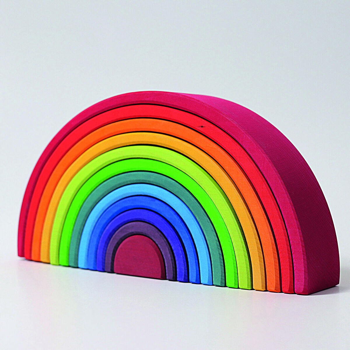 Ein buntes Holzspielzeug in Form eines Regenbogens. Es besteht aus mehreren ineinander stapelbaren halbrunden Teilen in den Farben Rot, Orange, Gelb, Grün, Blau und Lila. Das Spielzeug steht auf einer weißen Fläche.