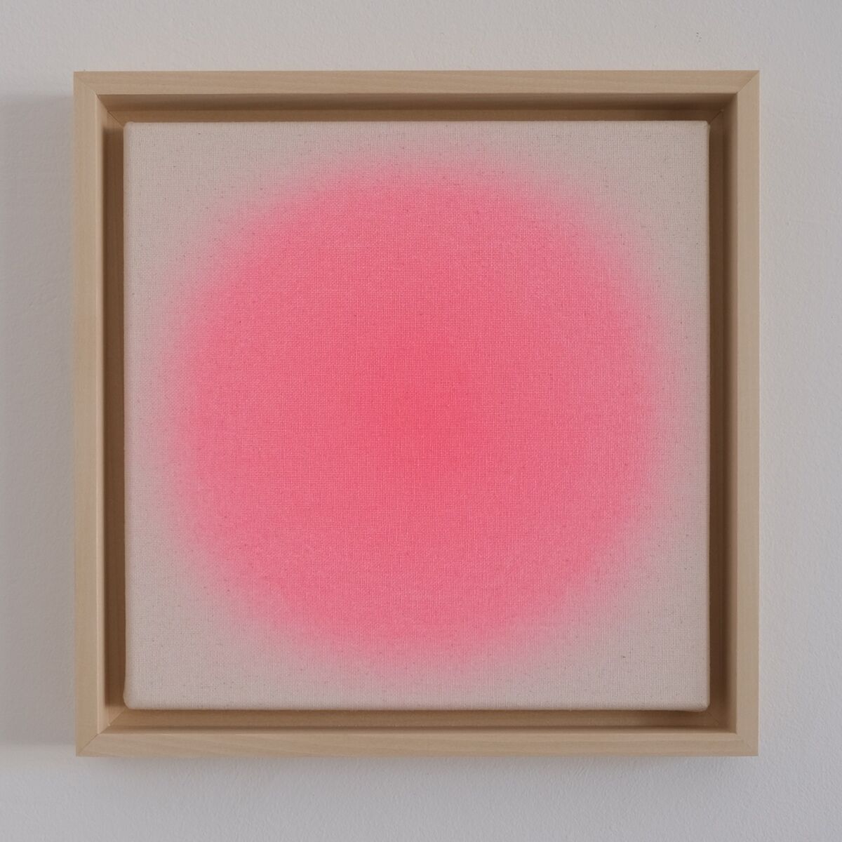 Quadratisches, minimalistisches Kunstwerk in einem hellen Holzrahmen. In der Mitte befindet sich ein leuchtend pinkfarbener, sanft auslaufender Farbverlauf auf naturweißer Leinwand vor neutralem Hintergrund. Das Bild vermittelt moderne, abstrakte Ästhetik.