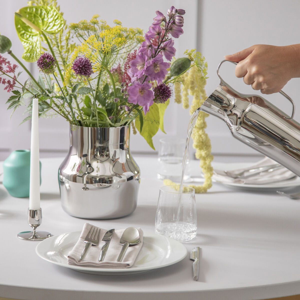 Ein eleganter Tisch mit dem Gense Besteckset 'Dorotea'. Das Besteck liegt auf weißen Tellern, mit einer modernen Vase und einer blühenden Blumendekoration in der Tischmitte. Ein Wasserkaraffe steht in der Nähe, bereit zum Eingießen.