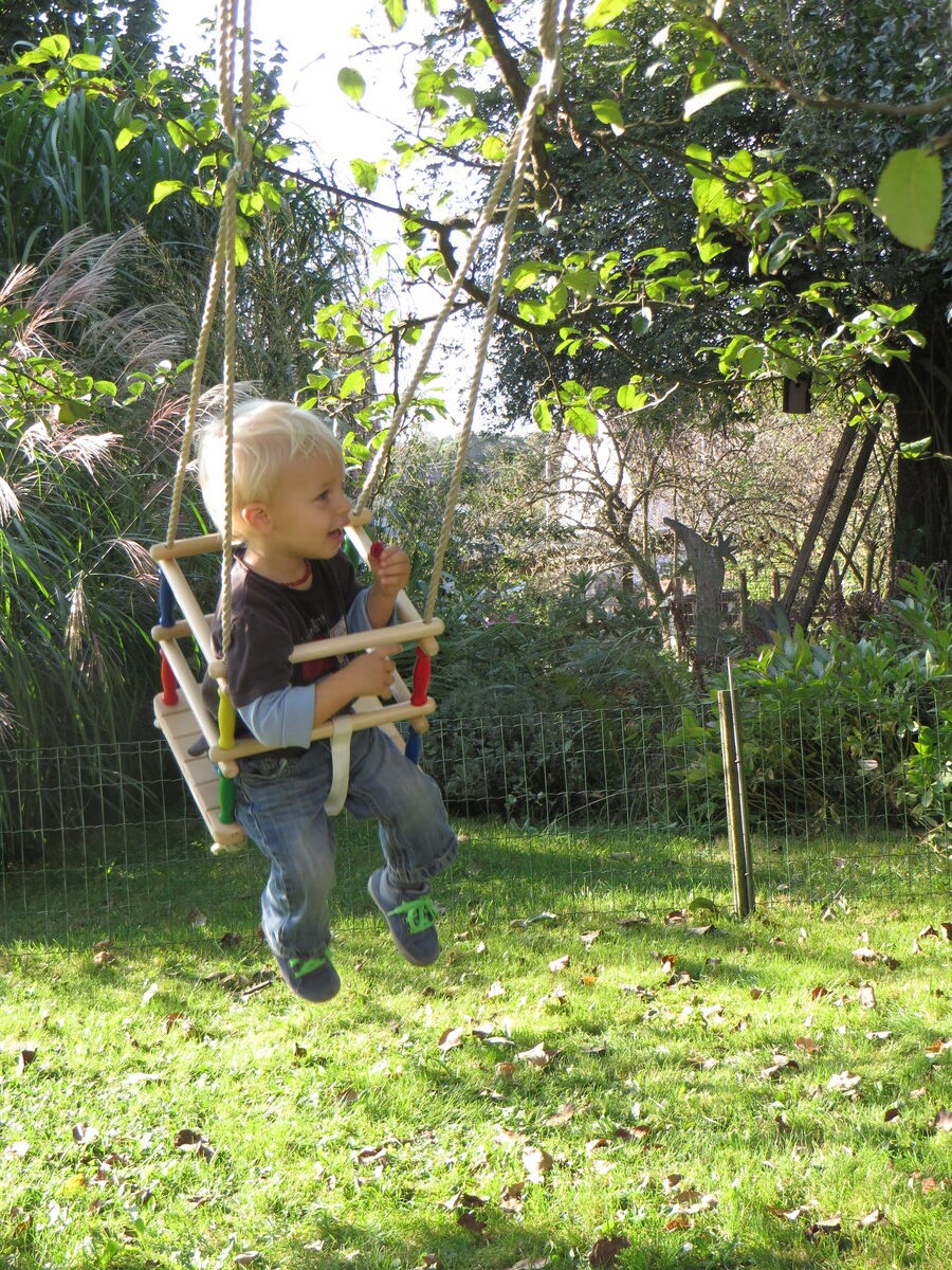 Ein kleines Kind sitzt in einer Schaukel aus Buchenholz im Garten. Rundherum sind grüne Bäume und Gras. Die Schaukel hat Seile, die an einem Baum befestigt sind.