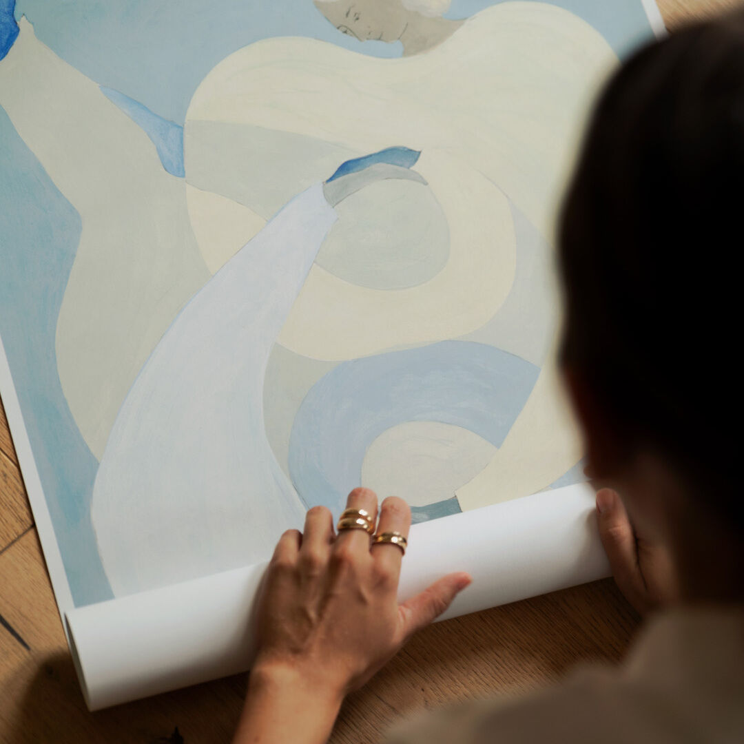 Eine Person mit mehreren Ringen an der Hand rollt ein Kunstplakat auf, das eine abstrakte Figur mit blauen und beigen Tönen zeigt. Das Plakat liegt auf einem Holzboden.