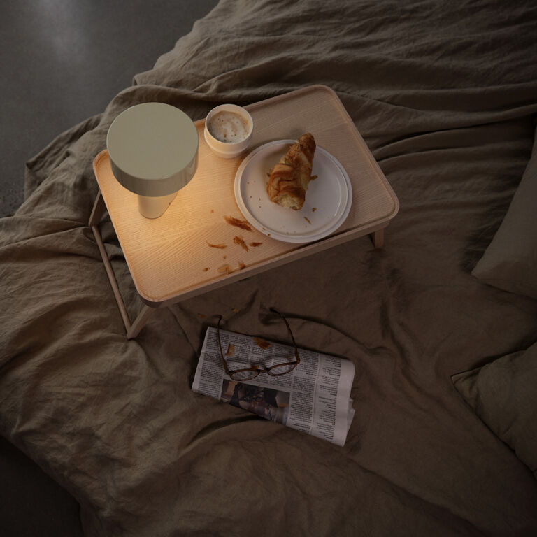 Eine tragbare, moderne Lampe namens 'Oblong' steht leuchtend auf einem hellen Holztablett, das auf einem ungemachten Bett liegt. Auf dem Tablett befinden sich ein französisches Croissant auf einem weißen Teller, eine kleine Schale mit Aufstrich, sowie Zeitungen und eine Brille daneben. Die Szene vermittelt eine gemütliche und entspannte Atmosphäre im Schlafzimmer.
