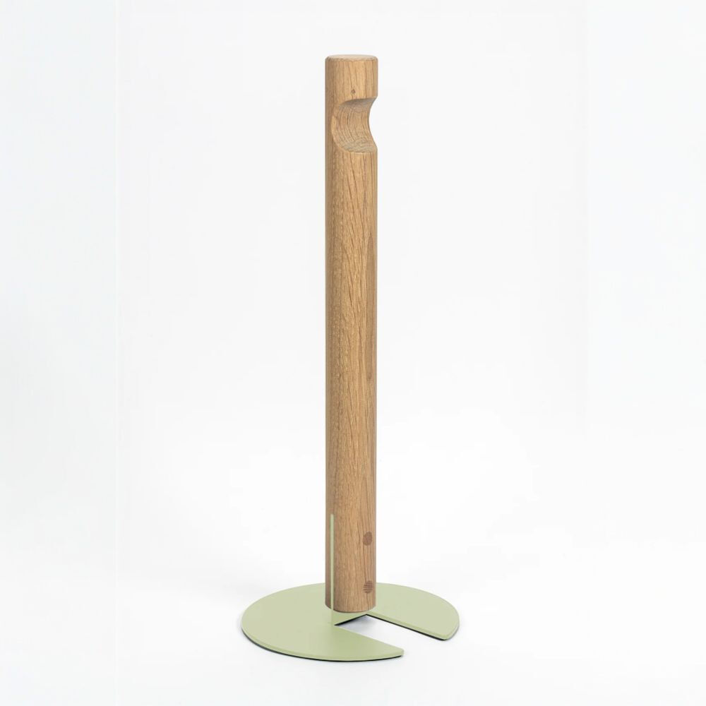 Freistehender Küchenrollenhalter GIVE von finbeaux vor weißem Hintergrund. Ein hoher, schlanker Stab aus hell gemasertem Holz mit eingeschnittener Griffmulde steht auf einem flachen, pistaziengrünen Metallfuß. Der runde Fuß ist vorn geöffnet und bildet zwei abgerundete Laschen; eine schmale graue Metallstrebe verbindet ihn mit dem Holzstab.
