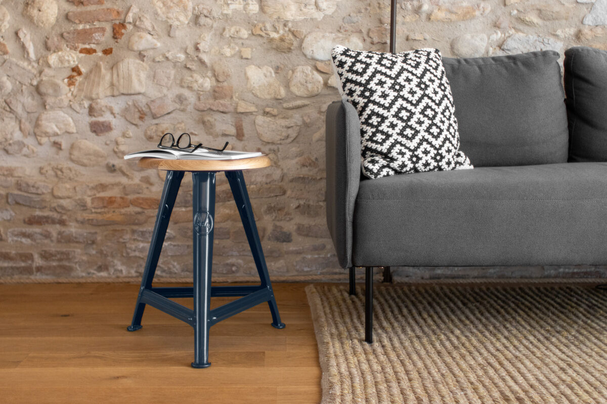 Ein stilvoller blauer Rowac-Schemel im Industrial-Design steht neben einem grauen Sofa und einem beigen Teppich. Auf dem Schemel liegt eine Brille und eine Zeitschrift, während ein gemustertes Kissen das Sofa ziert. Die Wand im Hintergrund besteht aus rustikalen Steinen.