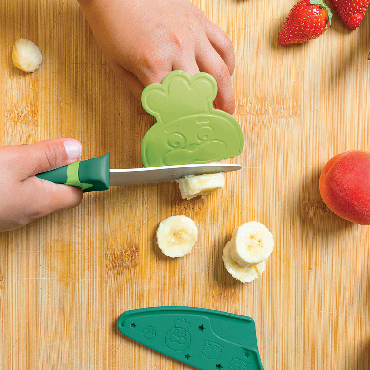 Ein kindersicheres, grünes Messer mit einem Tiergriff schneidet eine Banane auf einem hölzernen Schneidbrett, umgeben von frischen Erdbeeren und einem Pfirsich.