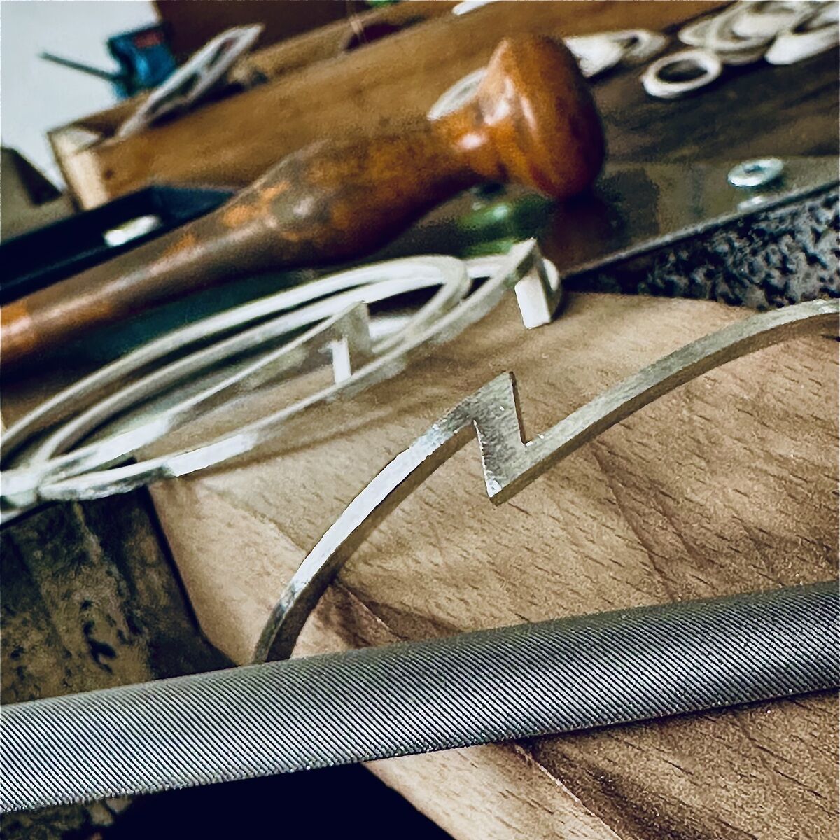 Detailaufnahme einer Werkbank mit einem silberfarbenen ZEIT Sonderedition Armreif »ZEIT« von HML Berlin, feingoldplattiert. Der Armreif liegt neben Werkzeugen wie einer Feile und einem Holzgriff, ringsherum weitere Schmuckelemente und Oberflächen aus Holz und Metall.
