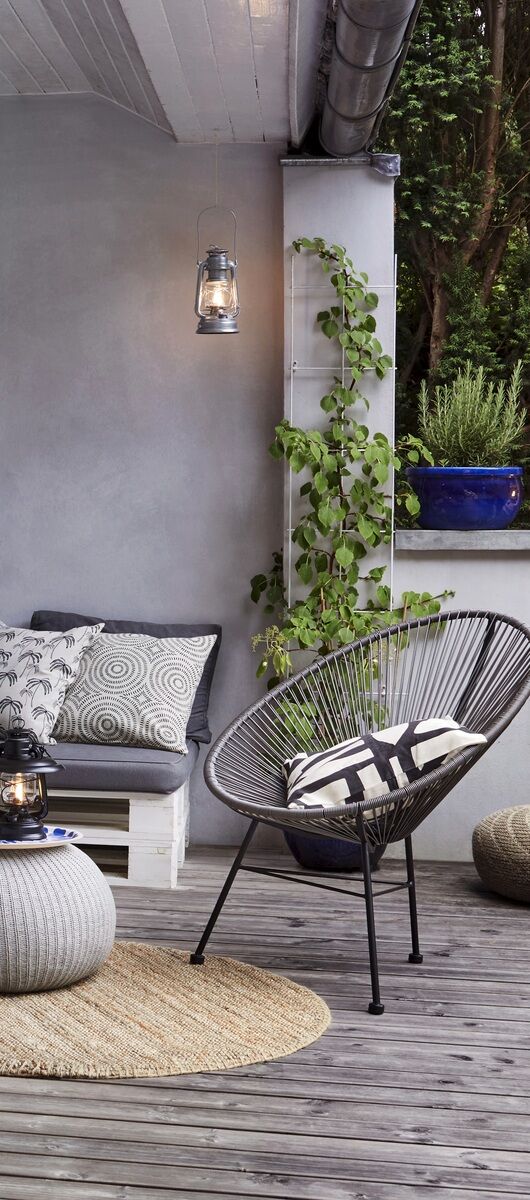 Eine moderne Terrasse mit gemütlicher Sitzecke. Eine LED-Laterne hängt an der Wand und eine weitere steht auf einem runden Hocker. Ein schwarzer, runder Rattansessel mit Kissen steht daneben. Kletterpflanzen wachsen an der Wand. Verschiedene Textilien und Pflanzen zieren den Raum. Der Boden ist aus Holz.