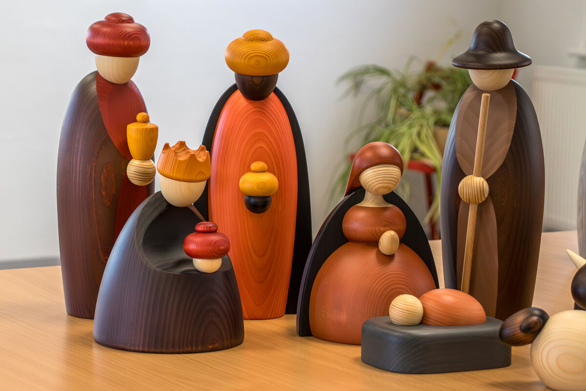 Fünf hölzerne Figuren mit runden Köpfen und farbigen Gewändern, eine liegende Figur mit Decke, alle auf einem Holztisch.