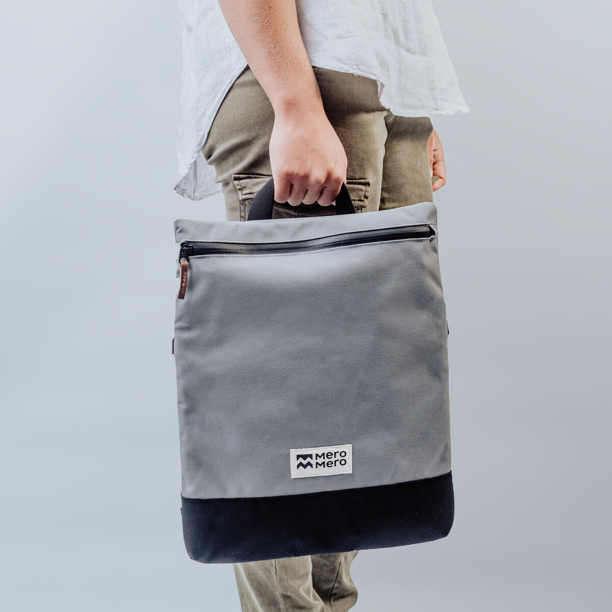 Eine Person, die eine hellgraue Stofftasche mit waagerechtem Reißverschluss und einem Etikett der Marke Mero Mero hält. Der untere Teil der Tasche ist dunkelgrau bis schwarz.