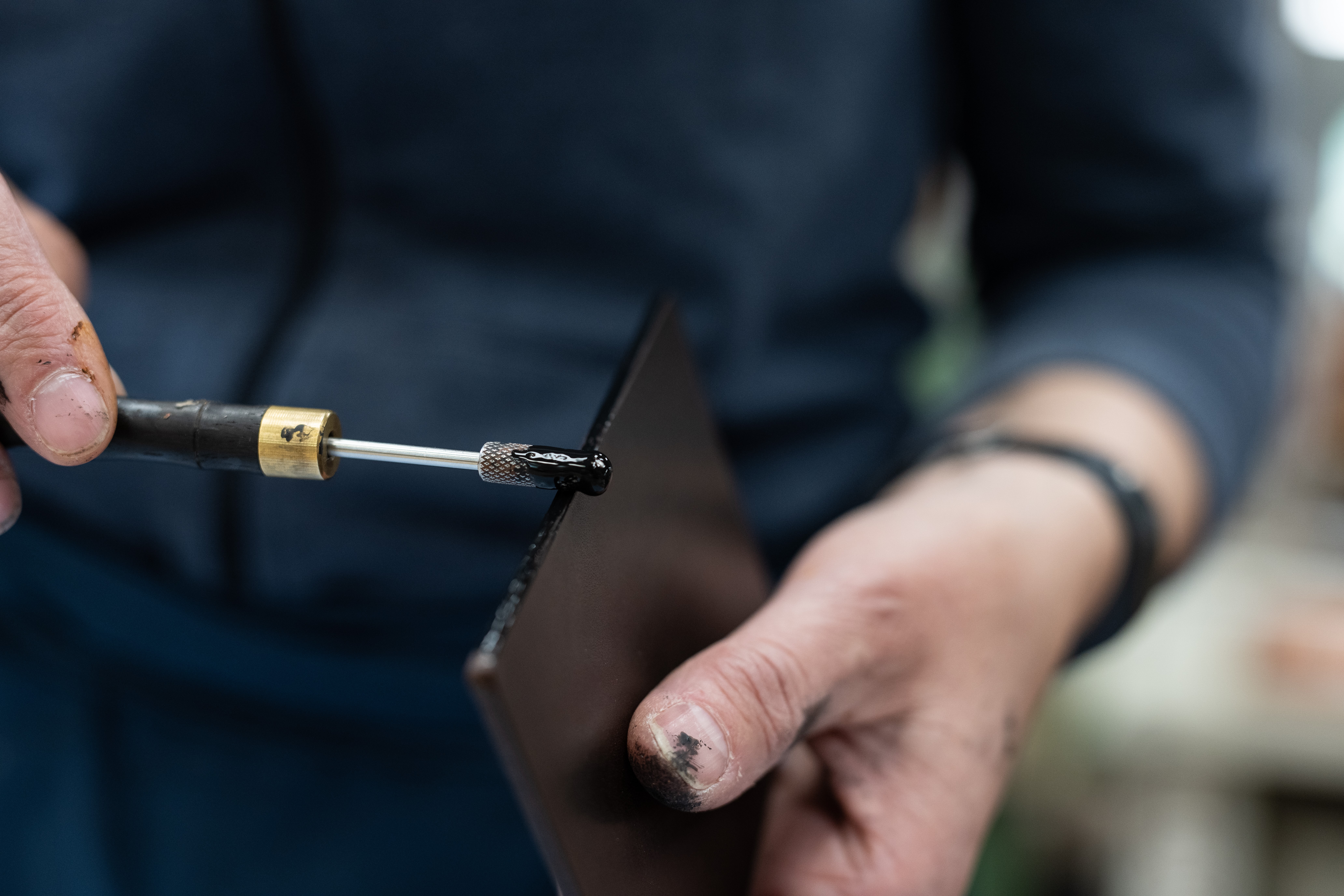 Nahaufnahme der Hand eines Handwerkers, der mit einem kleinen Werkzeug eine schwarze Flüssigkeit auf ein Metallblech aufträgt, wobei die handwerklichen Details deutlich werden.