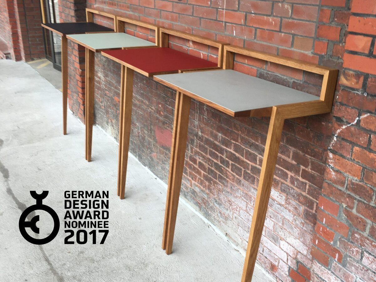 Drei Stehpulte der Marke »buckowski«, die entlang einer roten Backsteinwand aufgereiht sind. Die Stehpulte bestehen aus hölzernem Rahmen und Tischplatten in den Farben Schwarz, Grau, Rot und Weiß. Auf dem Bild ist auch das Logo des German Design Award Nominee 2017 zu sehen.
