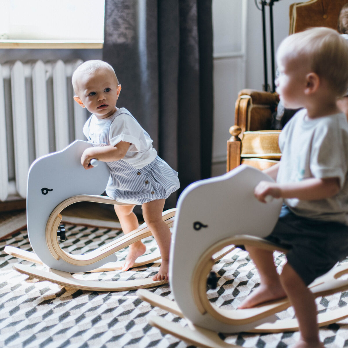 Zwei Kleinkinder schaukeln auf Elefant-förmigen Schaukelaufsätzen für Balance Bikes in einem hellen Kinderzimmer mit Teppich, während ein weiteres Kind auf einem Sessel im Hintergrund sitzt.
