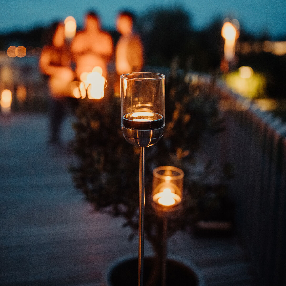 Mehrere Gravity Candles leuchten auf einer Terrasse bei Einbruch der Dunkelheit. Im Hintergrund sind unscharf drei Personen zu sehen, die sich unterhalten. Die Szene vermittelt eine warme, einladende Atmosphäre mit einem Hauch von Modernität.