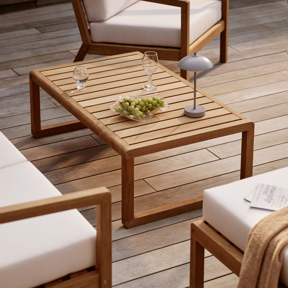 Helles Outdoor-Szenario mit einem rechteckigen Holztisch, auf dem eine tragbare, graue Lampe »Clam™« von Fritz Hansen, ein Weinglas, ein Wasserglas und Weintrauben stehen. Um den Tisch stehen moderne Sitzmöbel mit weißen Polstern, auf einem liegt eine beigefarbene Decke und eine Zeitung. Holzoptik-Dielenboden komplettiert das stilvolle skandinavische Ambiente.