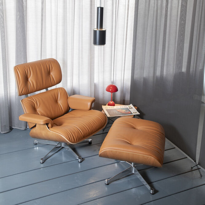 ZEIT-Sonderedition Lounge Chair und Ottoman. Ein brauner Ledersessel mit passendem Ottoman steht auf einem grauen Holzboden, umgeben von grauen Vorhängen. Ein kleiner roter Tisch mit Lampe und Zeitungen daneben.