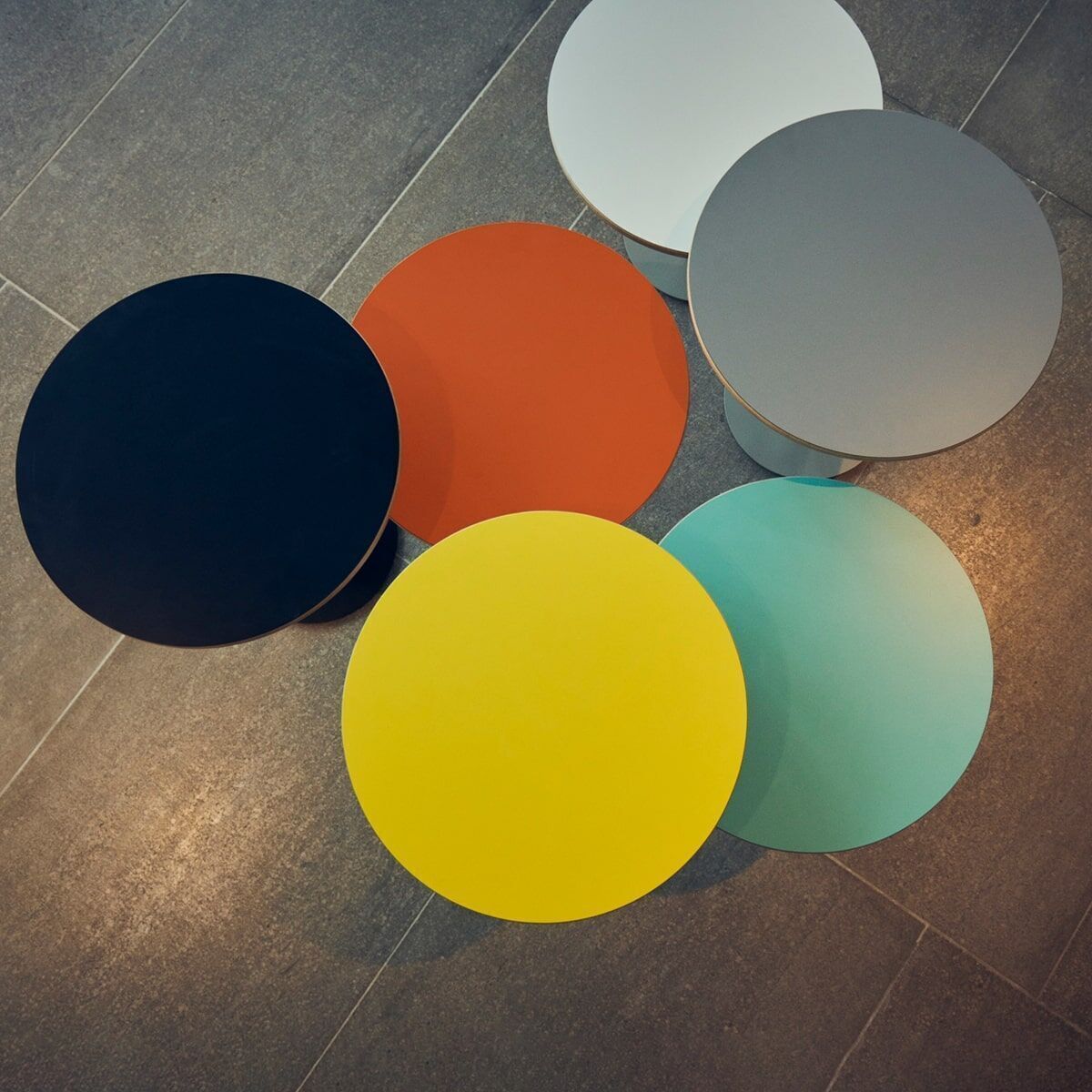 Fünf runde Beistelltische 'Panama' in verschiedenen Farben (Schwarz, Rot, Gelb, Türkis, Weiß, Grau) auf einem hellgrauen Boden angeordnet.
