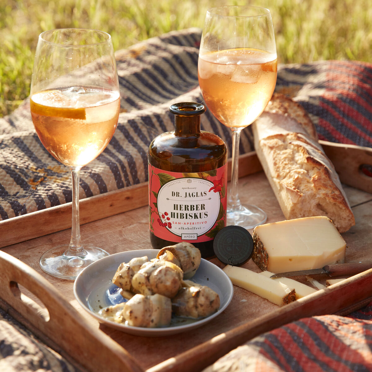 Ein Holztablett mit zwei Gläsern Rosé-Aperitif, einer kleinen Flasche »Herber Hibiskus« Aperitif, Käsestücken, Brot und Pilzen, alles auf einer Picknickdecke im Freien.