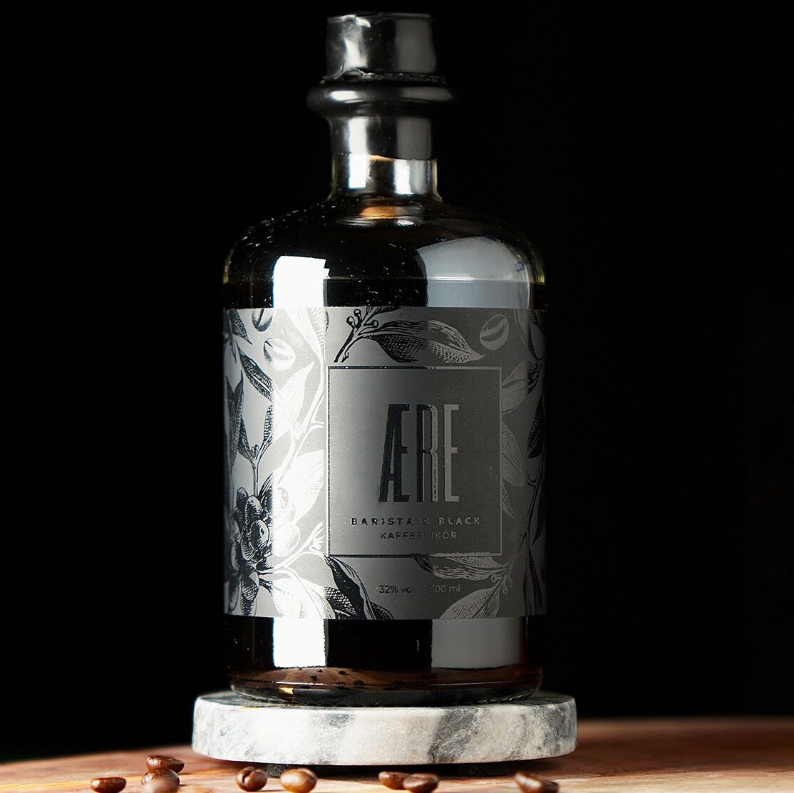 Eine edle schwarze Flasche AERE Barista's Black Kaffeelikör steht auf einem runden Untersetzer aus Marmor, umgeben von Kaffeebohnen.