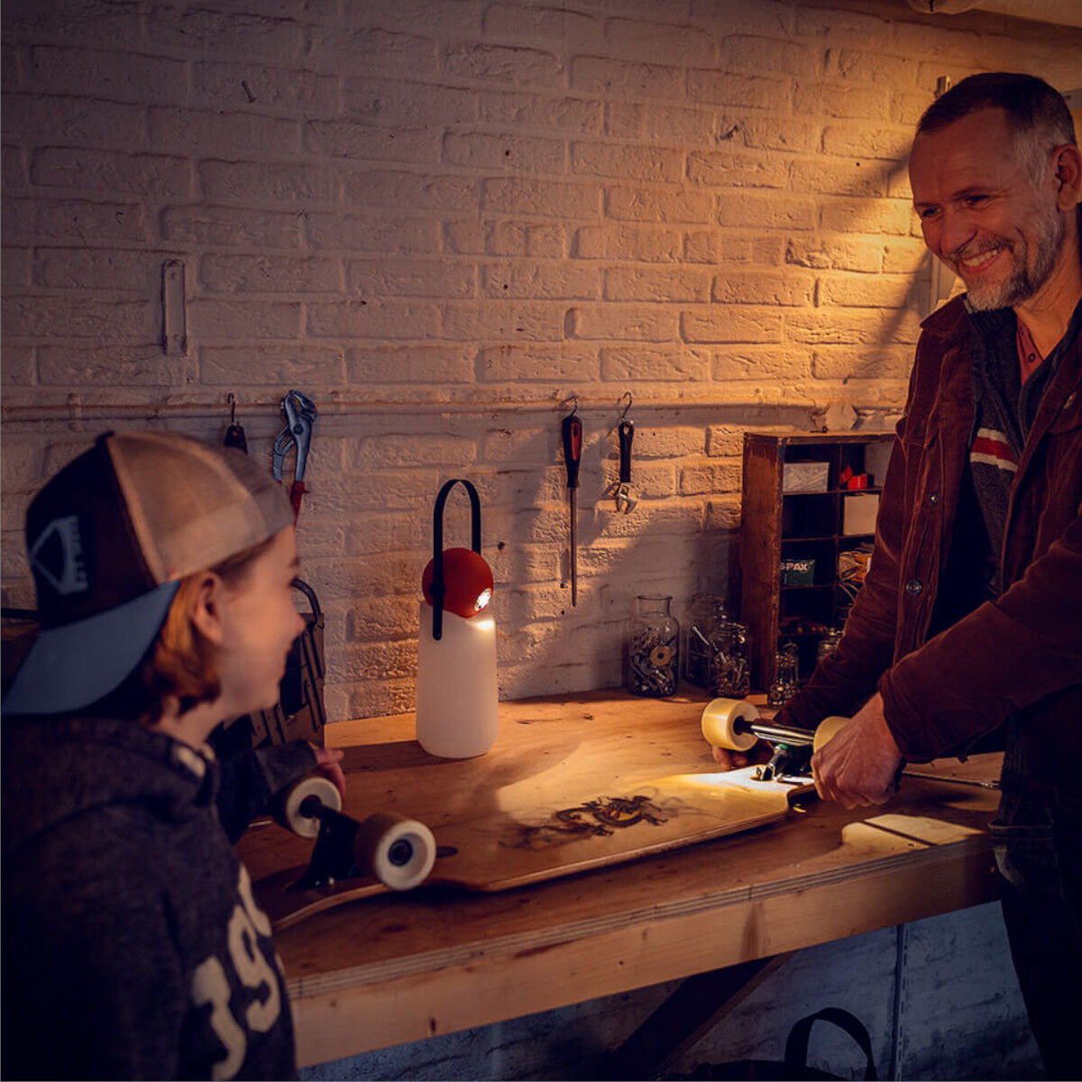 In einer gemütlichen Holzwerkstatt stehen ein Vater und sein Sohn an einer Werkbank. Die »Guidelight« Lampe leuchtet den Arbeitsplatz stimmungsvoll aus. Verschiedene Werkzeuge hängen an der Wand, während auf der Werkbank eine Skateboard-Achse bearbeitet wird.