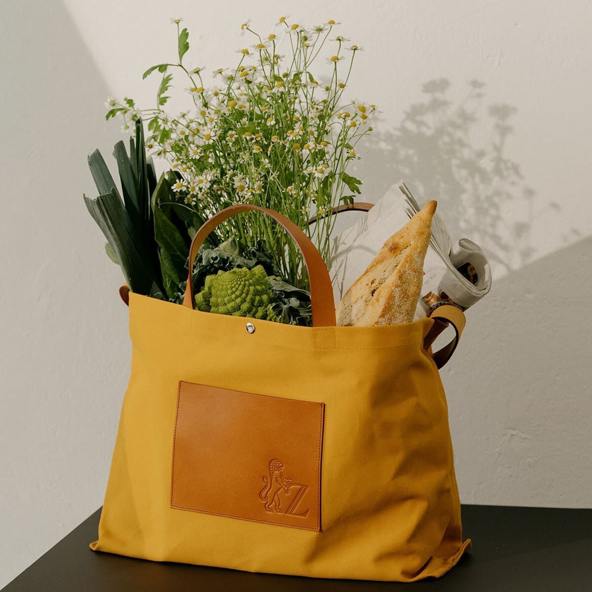 Die Jubiläums-Markttasche ist eine gelbe Einkaufstasche mit braunen Griffen und einer braunen Tasche auf der Vorderseite. Sie ist gefüllt mit frischen Blumen, einem Baguette und Gemüse und steht vor einem hellen Hintergrund.