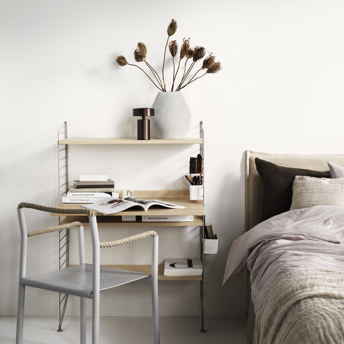 Ein minimalistischer Arbeitsplatz bestehend aus einem schlanken, modularen String-Möbelsystem mit Regalen, einer Arbeitsfläche und einer Metallstuhl mit einem Kissen. Der Schreibtisch ist in einem modernen, schlichten Design, das perfekt zu einem hellen, gut organisierten Raum passt. Neben dem Arbeitsplatz befindet sich ein schlichtes Bett. Auf dem Schreibtisch stehen Bücher, Schreibutensilien und eine Vase mit getrockneten Blumen. Die Farben im Bild sind Weiß, Beige, Grautöne und verschiedene Holztöne.