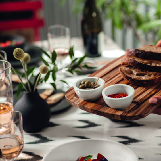 Ein schön gedeckter Tisch mit einem hölzernen Schneide- und Servierbrett, das verschiedene Brotscheiben und Schalen mit Oliven und anderen Snacks präsentiert. Im Hintergrund sind eine Blumenvase und eine Flasche zu sehen.