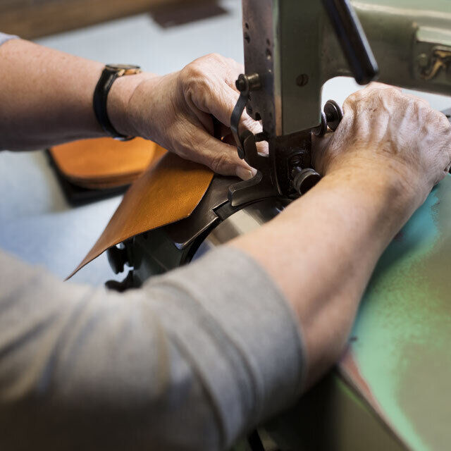 Eine Person arbeitet an einem Lederstück in einer Werkstatt, wahrscheinlich um ein Stift- oder Kosmetik-Etui zu fertigen. Man sieht Hände, die das Leder unter einer Nähmaschine führen. Die Farben des Bildes sind vorwiegend Braun, Beige, Silber und Grün.