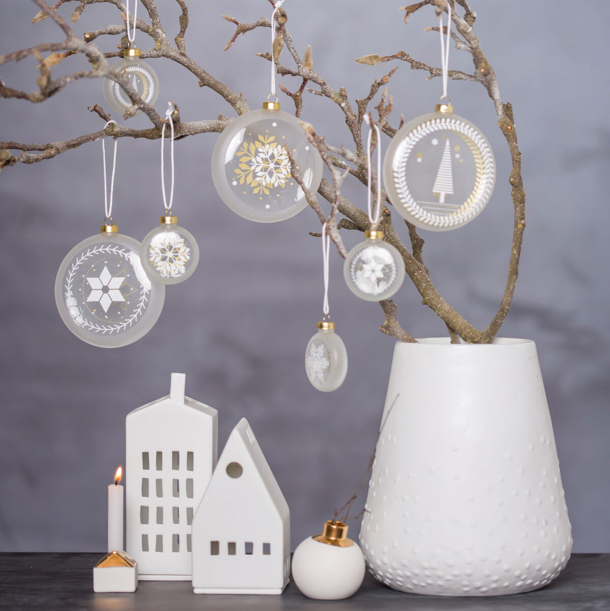Zwei weiße Keramikhäuser stehen auf einem Tisch neben einer weißen Vase mit kahlen Zweigen, die mit durchsichtigen Ornamenten und Schneeflockenmotiven geschmückt sind. Ein kleines Teelicht leuchtet neben den Häusern.