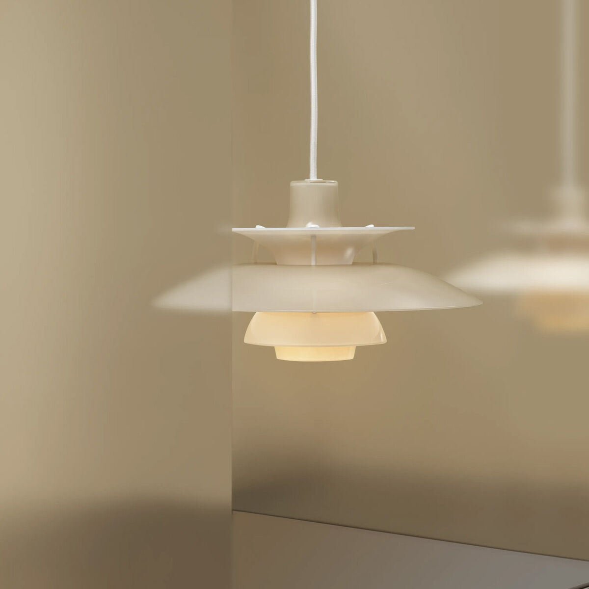 Die Hängeleuchte »PH 5 320« von Louis Poulsen in opal-beige hängt vor einem beige Hintergrund. Die charakteristischen, übereinander angeordneten Schirme sorgen für blendfreies, sanftes Licht. Ein Spiegel reflektiert die Leuchte und verstärkt den eleganten Effekt.