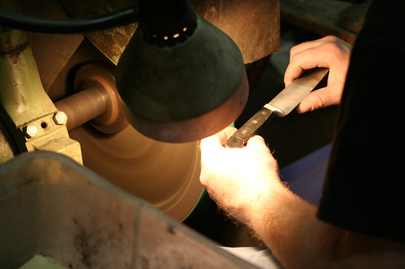 Ein Handwerker schärft ein Schinkenmesser mit einem Olivenholzgriff unter einer Lampe an einer Schleifmaschine. Das Bild zeigt den Fokus auf die Hände und das Messer.
