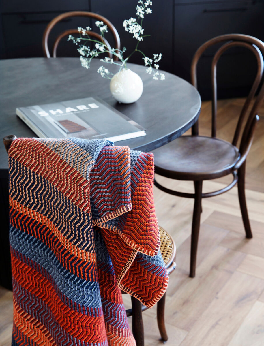 Eine Lammwolldecke mit orange, braunen und blauen Zickzackmustern hängt über einem Holzstuhl. Im Hintergrund steht ein runder Tisch mit einer Vase weißer Blumen und Lesematerial. Der Boden ist aus Holzparkett.