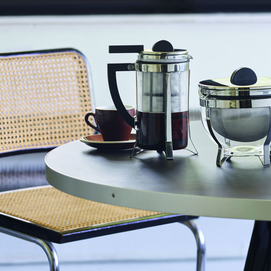 Ein Kaffeebereiter im Bauhaus-Stil steht auf einem runden Tisch neben einer Tasse und einem weiteren Behälter. Im Hintergrund ist ein Stuhl mit Korbgeflecht und ein Fenster mit Jalousien zu sehen.