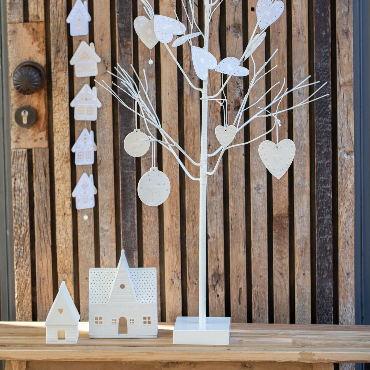 Ein dekoratives weißes Lichthaus »Lebkuchen Groß« auf einem Holztisch vor einer Holzlattenwand. Neben dem Lichthaus befindet sich ein weißer Weihnachtsbaum mit herzförmigen Ornamenten und anderen weihnachtlichen Dekorationen.