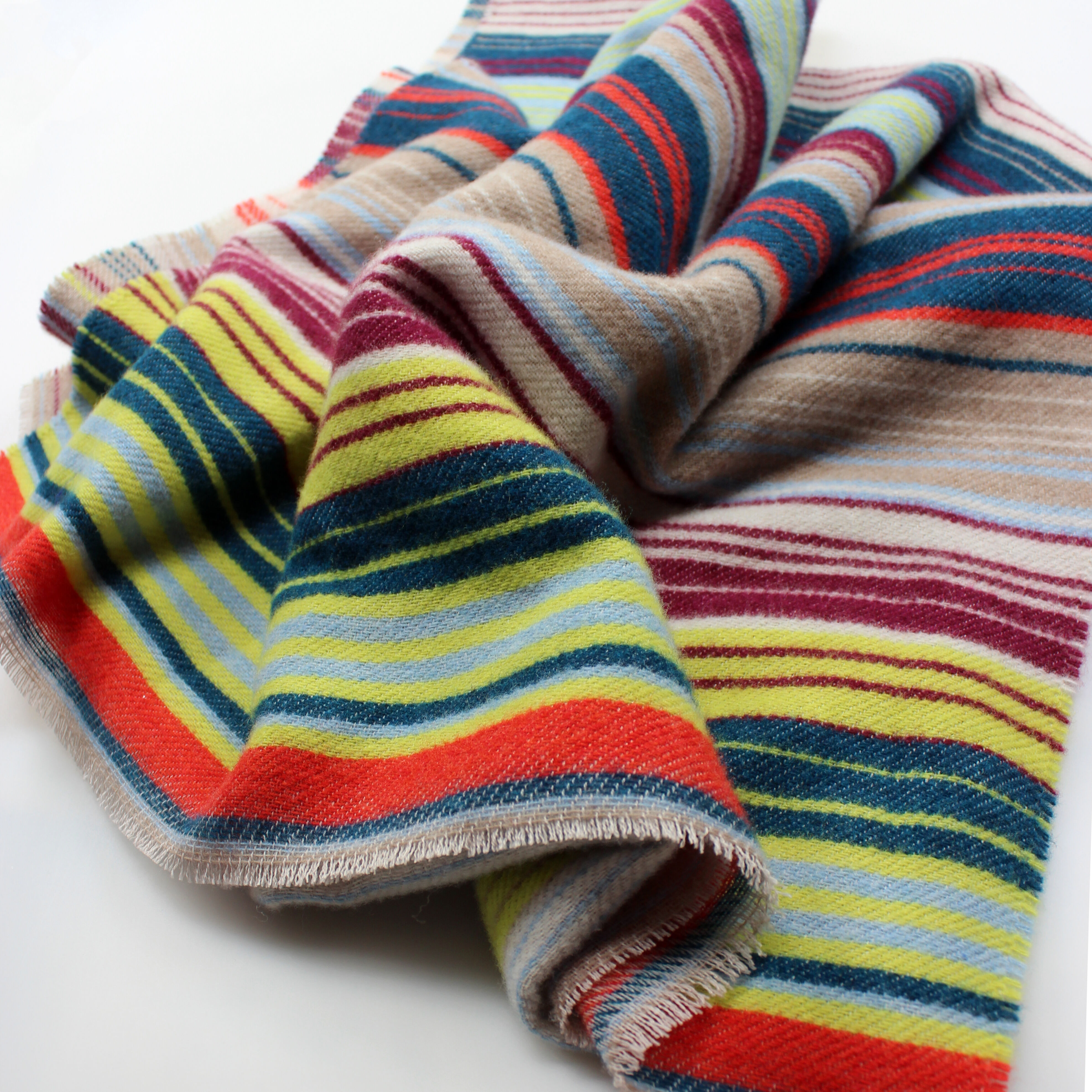Nahaufnahme eines weichen Wollschals mit buntem Streifenmuster in Rot, Gelb, Grün, Blau, Beige, Braun, Weiß und Orange. Der Schal liegt locker drapiert auf einer weißen Oberfläche.
