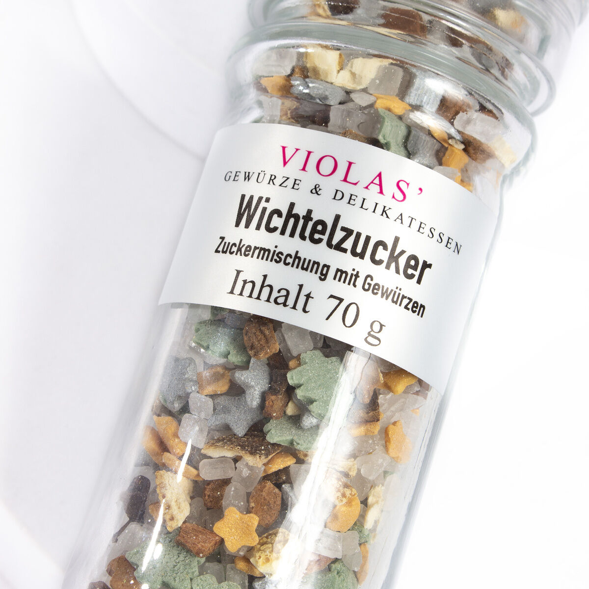 Ein Glasbehälter mit der Beschriftung 'VIOLAS' Gewürze & Delikatessen Wichtelzucker Zuckermischung mit Gewürzen'. Der Inhalt besteht aus gemischten Zuckerstreuseln in den Farben Gold, Silber, Grün, Braun und Weiß in Stern- und Granulatform.