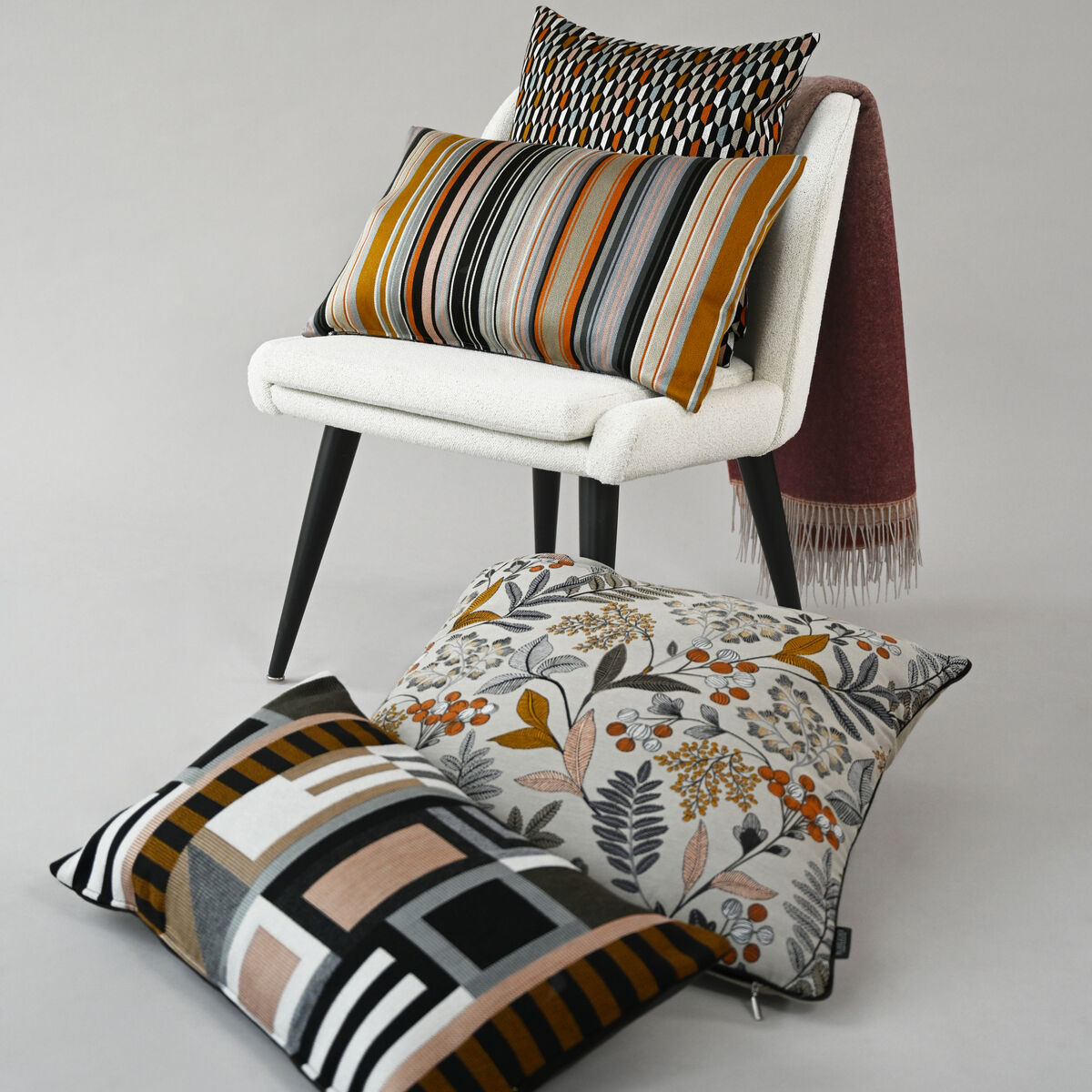 Ein Set von dekorativen Kissen auf einem weißen Sessel. Die Kissen haben verschiedene Muster, darunter Streifen, geometrische Designs und florale Elemente in Farben wie Orange, Grau, Schwarz und Weiß. Die Szene vermittelt einen modernen, gemütlichen Eindruck.