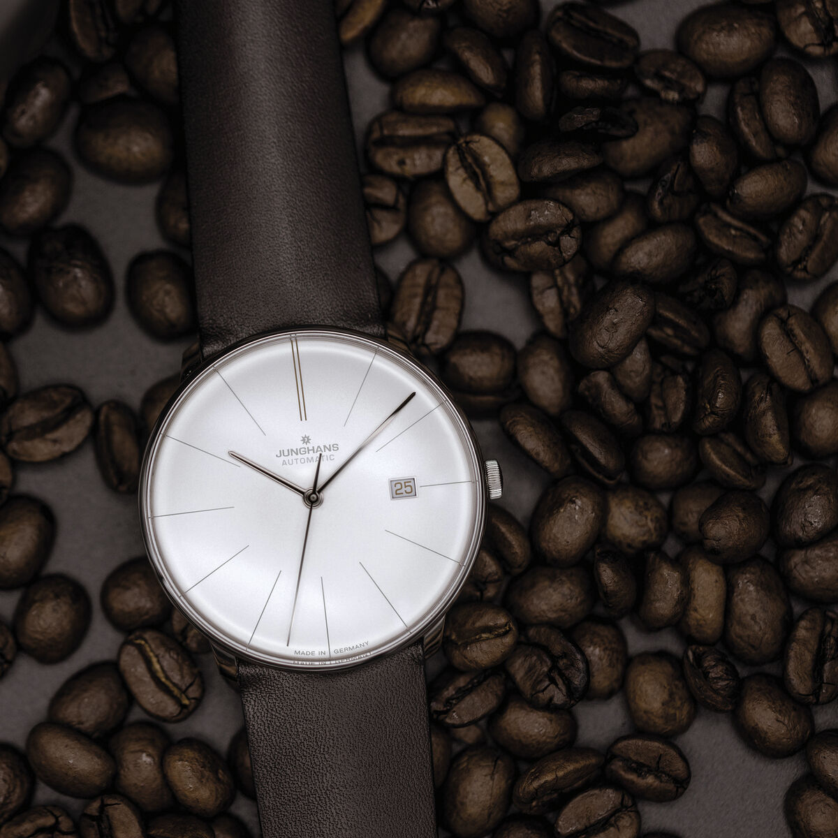 Die ZEIT-Sonderedition »Meister fein Automatic« von Junghans ist eine elegante Armbanduhr mit einem minimalistischen Design. Das Zifferblatt ist weiß, das Lederarmband ist in Schwarz gehalten. Die Uhr liegt auf einem Bett aus Kaffeebohnen.