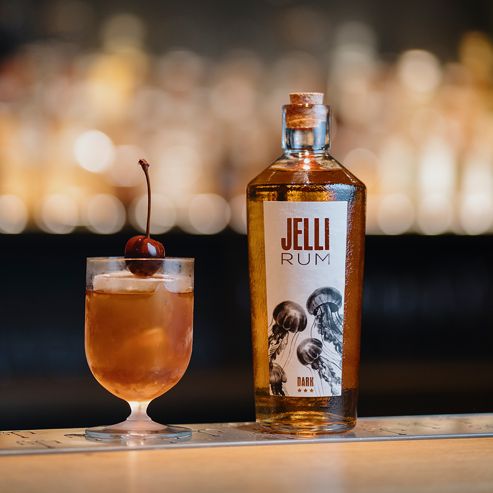 Flasche JELLI Rum Dark mit Korkverschluss neben Cocktailglas mit Eiswürfeln und Cocktailkirsche auf hellem Tresen.