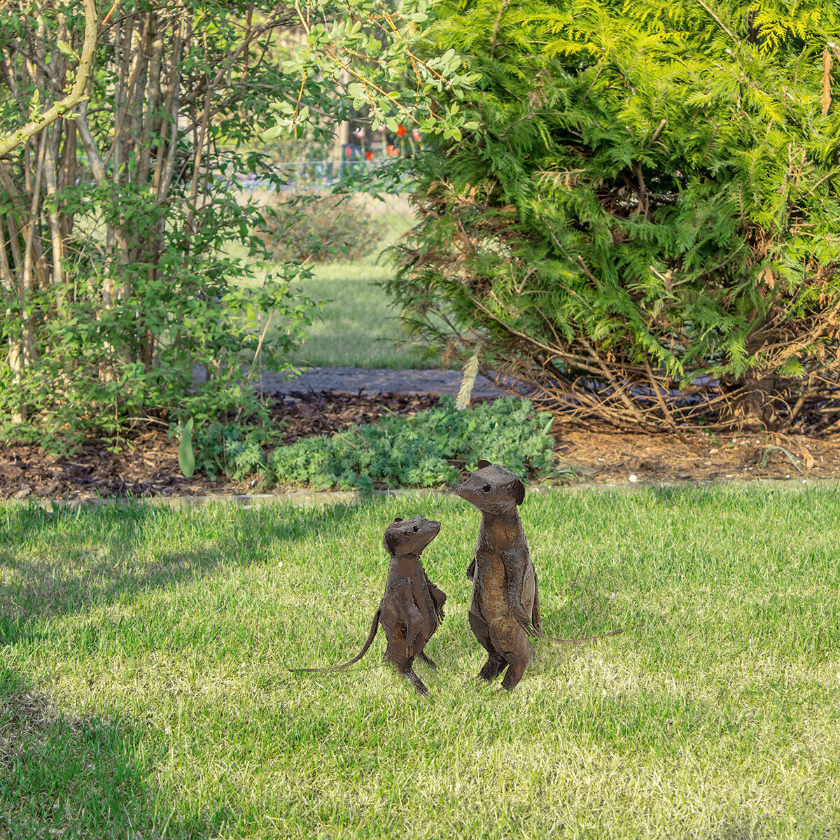 Zwei Gartenfiguren in Form von Erdmännchen stehen auf grünem Rasen vor einem Hintergrund aus Büschen und Pflanzen. Eine Figur ist größer und die andere kleiner, beide schauen in dieselbe Richtung.