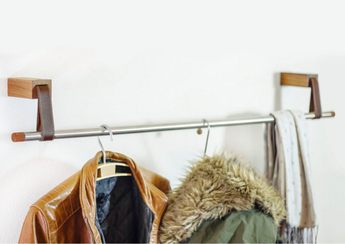 Ein minimalistisches Garderobensystem 'Ansa' aus Eiche, montiert an einer weißen Wand. Die Halterungen bestehen aus Stahl und Holz. Daran hängen eine Lederjacke, ein Mantel mit Fellkapuze und ein Schal.