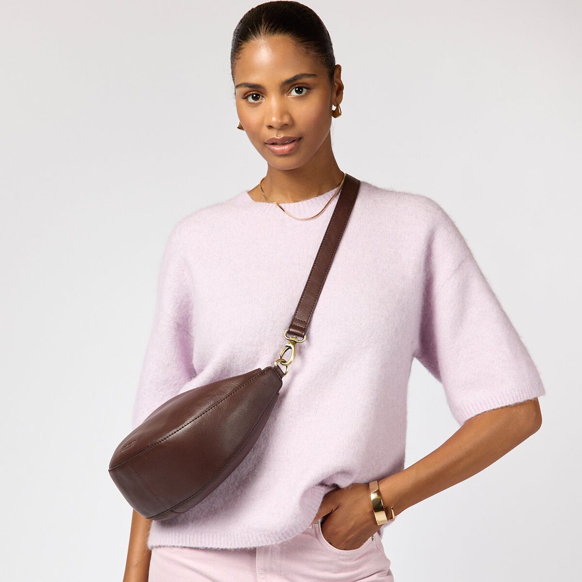 Eine Frau trägt eine Crossbody-Bag »Leo« aus schokoladenbraunem, offenporigem Leder von O My Bag über eine pastell-lavendelfarbene Strickpullover. Die Tasche liegt nah am Körper, der Gurt ist verstellbar, das Design ist schlicht und edel mit goldfarbenen Beschlägen.