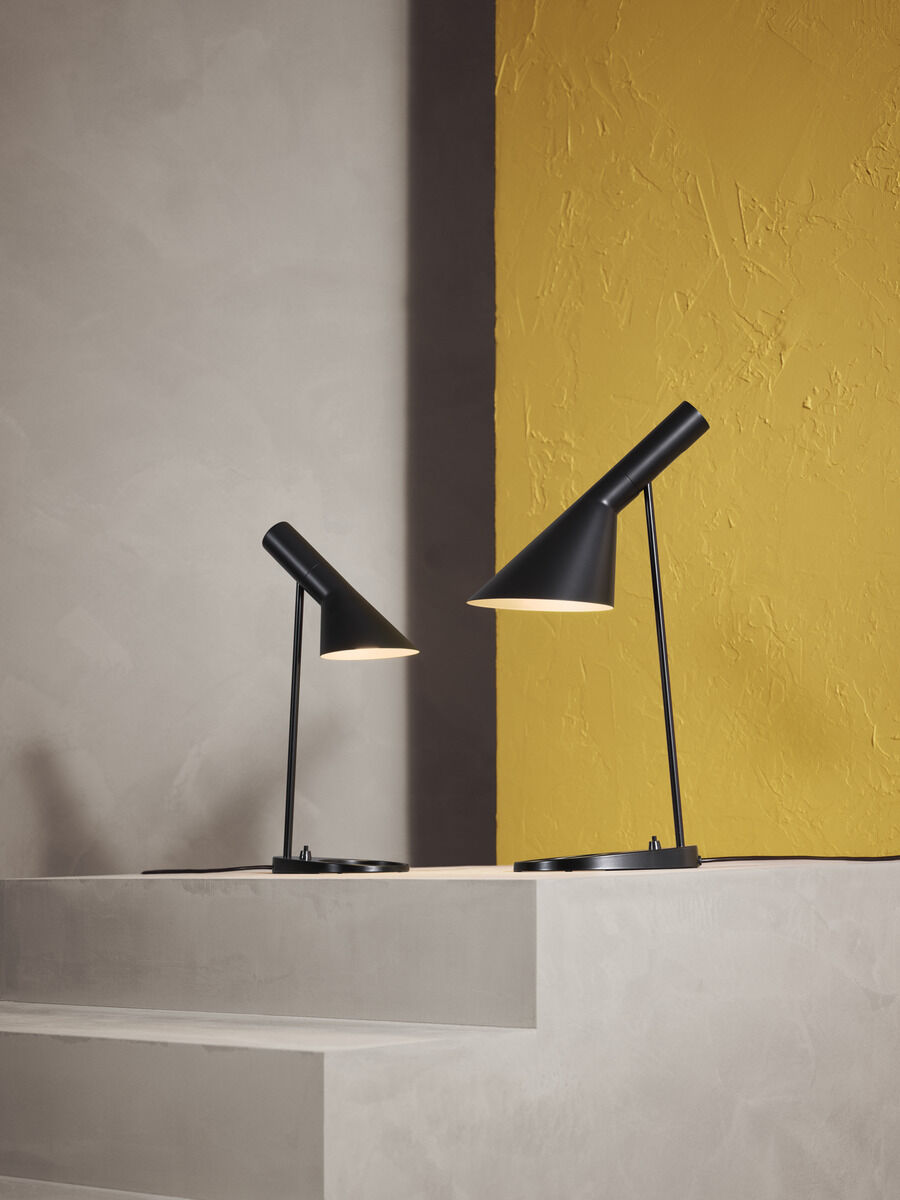 Zwei schwarze Tischlampen des Modells AJ Table Mini stehen auf gestaffelten weißen Plattformen vor einer zweifarbigen Wand in gelb und grau.