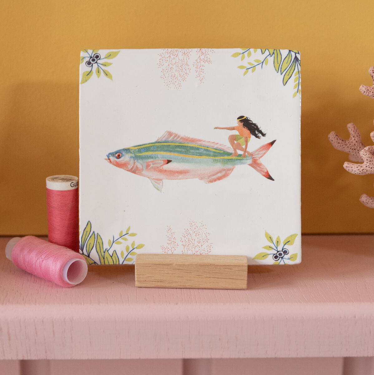 Keramikfliese »Auf den Wellen reiten« mit einer handgemalten Illustration eines Fisches und einer kleinen Person, die darauf reitet. Die Fliese hat einen weißen Hintergrund mit bunten Akzenten und floralen Mustern an den Ecken. Perfekt für Badezimmer- oder Küchendekor.