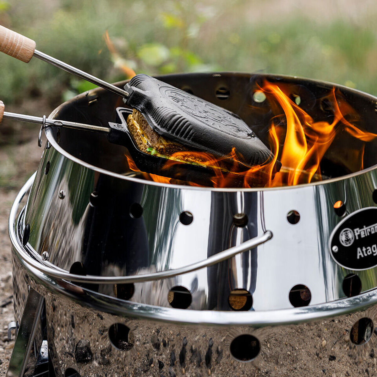 Ein Sandwicheisen mit Holzgriffen wird über einer Feuerstelle auf einem silbernen Grill erhitzt. Flammen lodern um das Eisen, während es ein Sandwich röstet. Im Hintergrund ist eine grüne Grasfläche zu sehen.