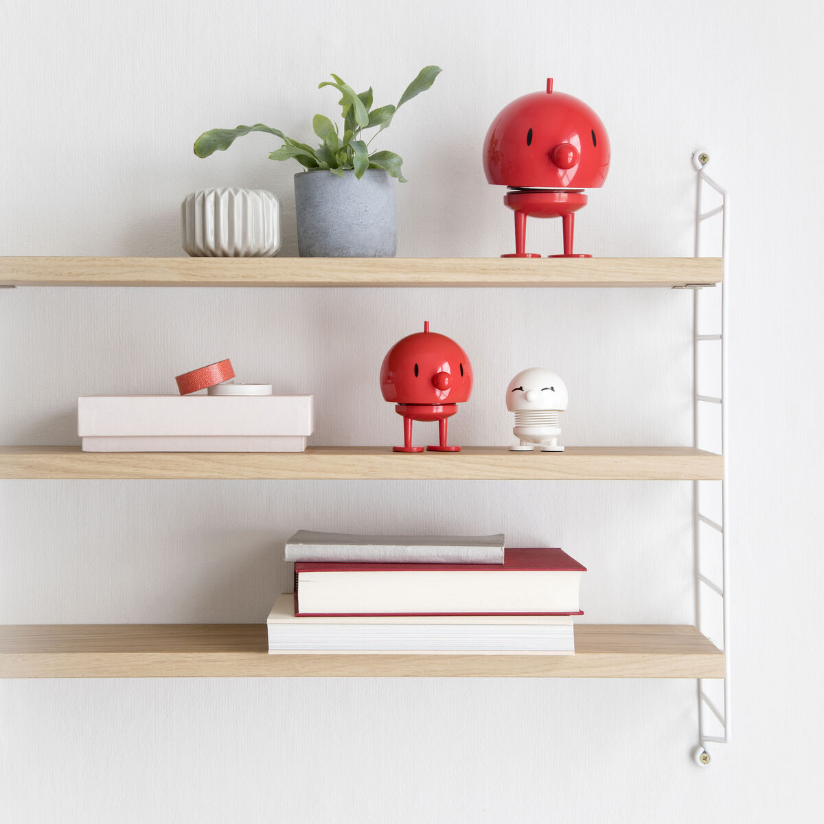 Ein minimalistisches Holzregal an einer weißen Wand mit Hoptimist Bumble Figuren in der Farbe Rot, Pflanzen und Büchern als Dekoration.