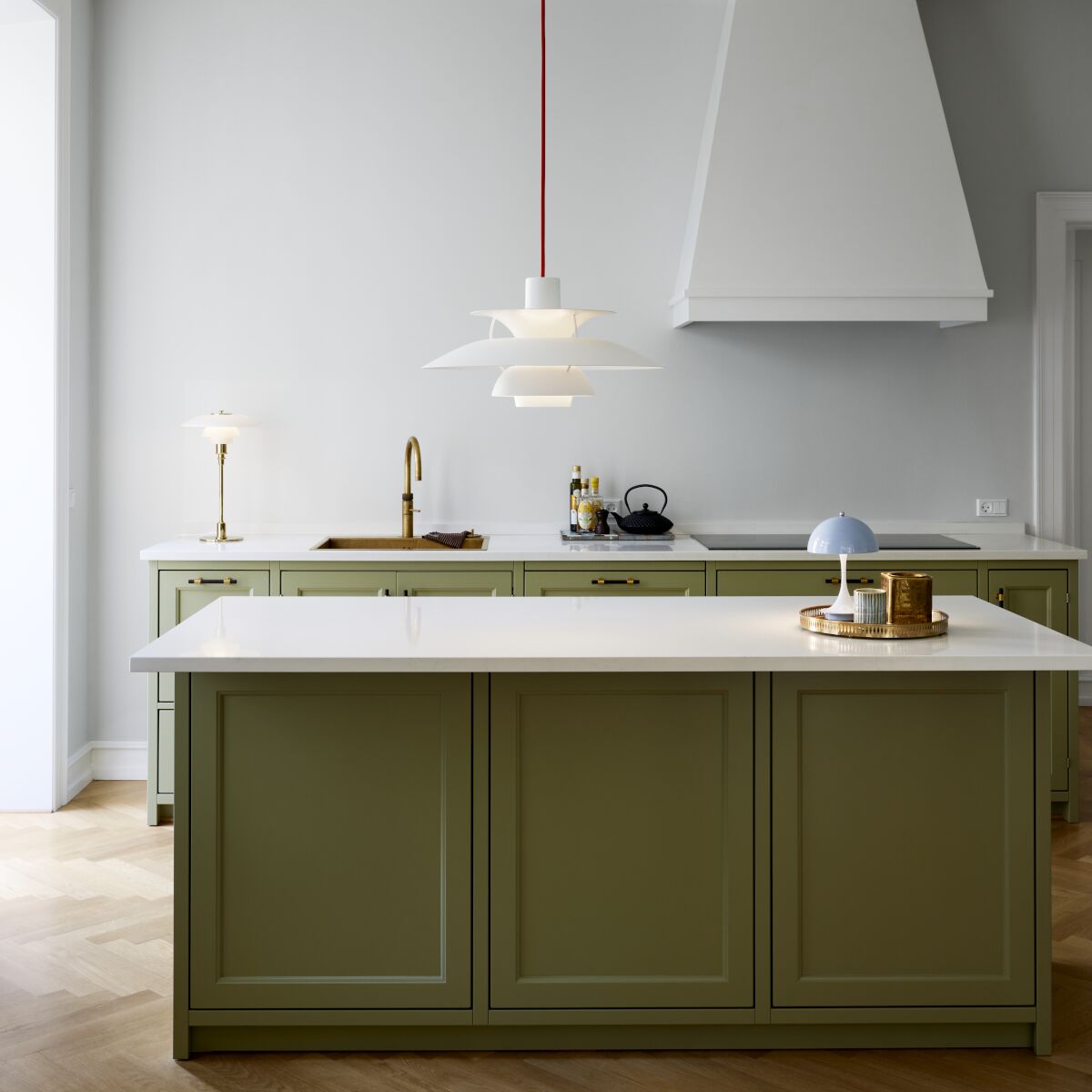 Zeitgenössische Küche mit olivgrüner Kochinsel und moderner Pendelleuchte der ZEIT-Sonderedition PH5 Monochrome in Weiß und Rot, hängend über einer hellen Küchenarbeitsplatte. Im Hintergrund befindet sich eine weiße Dunstabzugshaube, eine dezente Tischleuchte, goldene Accessoires und helle Parkettböden. Das Bild vermittelt ein Gefühl von modernem, stilvollem Wohnen.