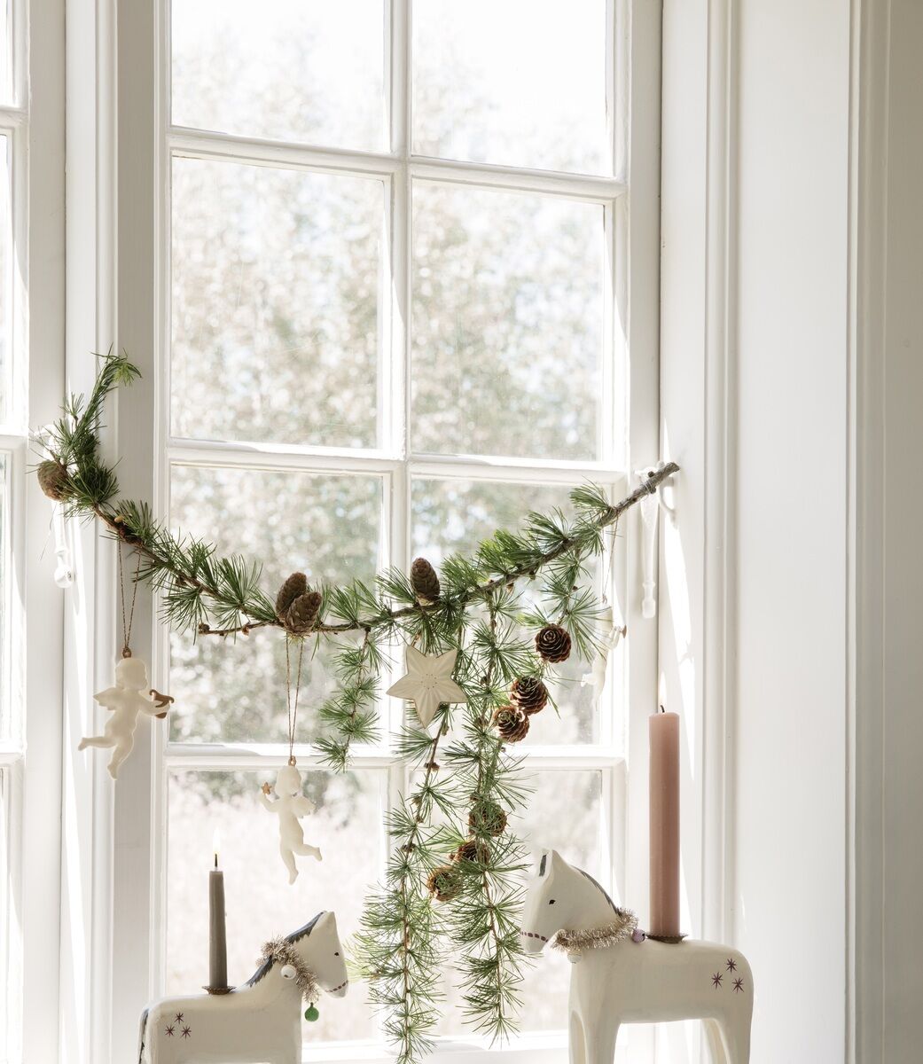 Set von Weihnachtsanhängern namens 'Heilige Nacht', aufgehängt an einem Ast aus grünen Tannenzweigen mit Kieferzapfen, weißen Engeln und Sternen. Darunter stehen Kerzenhalter in Form von weißen Pferden vor einem Fenster. Die Szene wird von natürlichem Tageslicht beleuchtet.