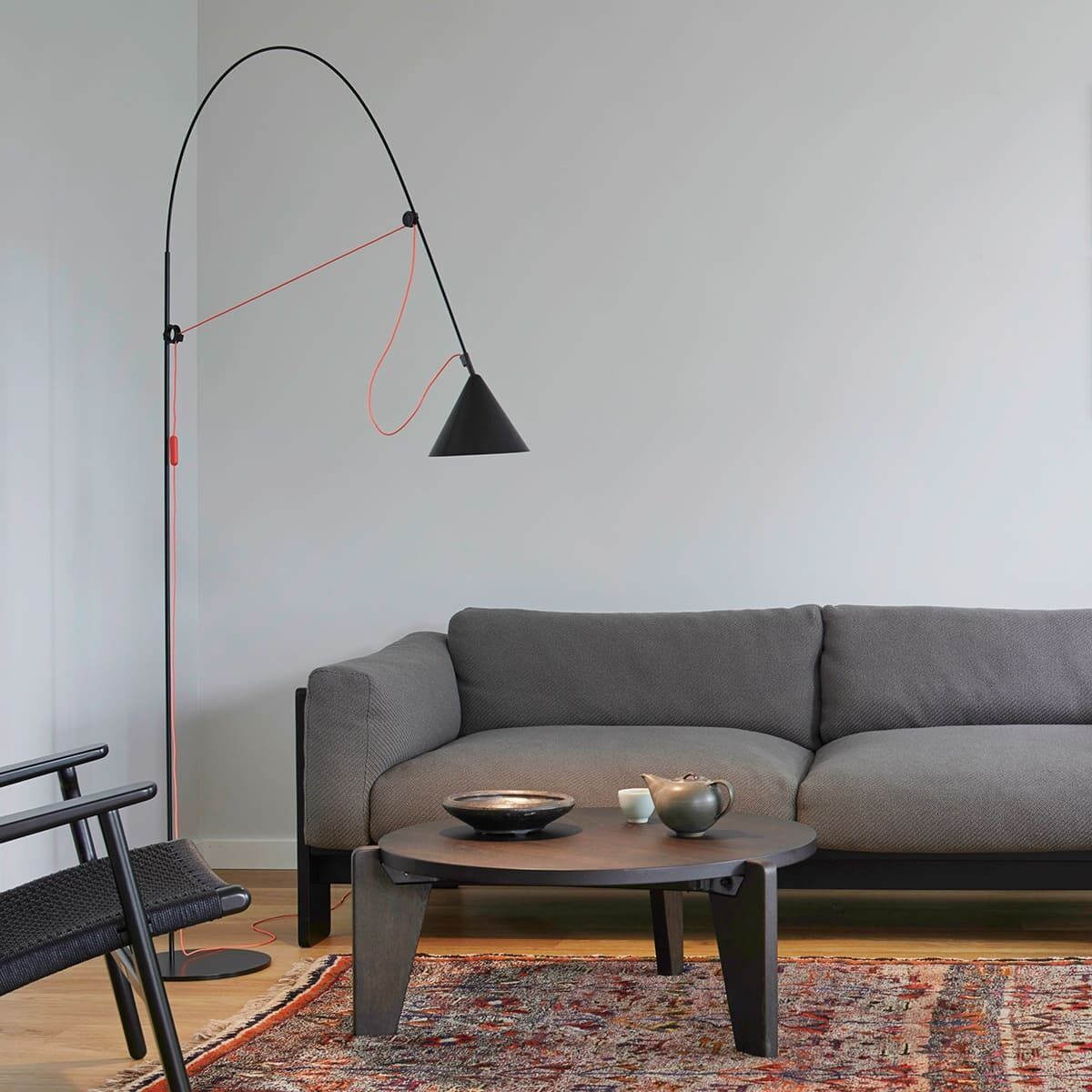Ein modernes Wohnzimmer mit einer stilvollen Stehlampe „AYNO“, die eine schlanke, gebogene schwarze Struktur und einen kegelförmigen Lampenschirm hat. Neben einem grauen Sofa und vor einem bunten Teppich steht ein runder schwarzer Couchtisch.